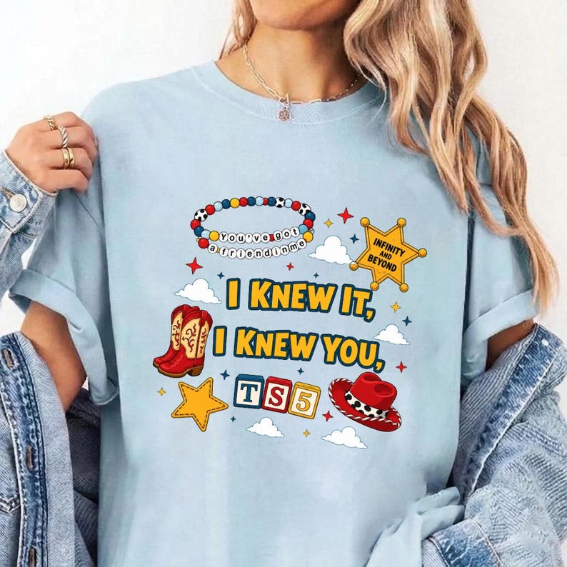 May include: A light blue t-shirt featuring a graphic print with the text I KNEW IT, I KNEW YOU, TS5, and INFINITY AND BEYOND. The design includes a colorful beaded bracelet, a yellow sheriff star, red cowboy boots, a red cowboy hat, and clouds.