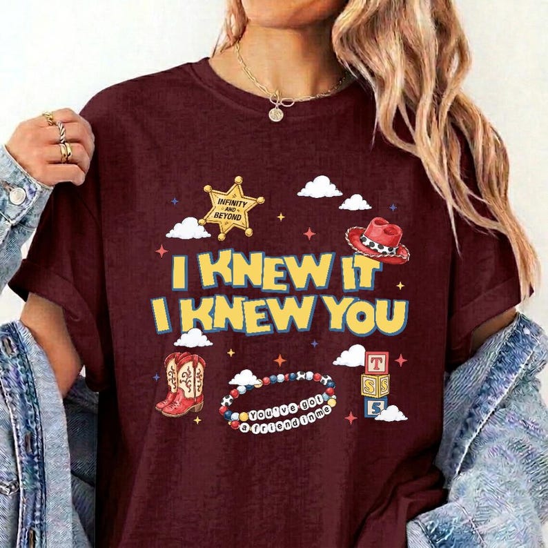 Puede incluir: Una camiseta granate con un estampado gr&aacute;fico que incluye el texto I KNEW IT I KNEW YOU, una estrella de sheriff dorada con el texto INFINITY AND BEYOND, un sombrero de vaquero rojo, botas vaqueras rojas, una pulsera de cuentas y tres cubos.