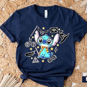 Puede incluir: Una camiseta azul marino con un personaje azul Stitch que lleva una bufanda y gafas. La camiseta presenta un diseño gráfico con varios elementos temáticos de Harry Potter, incluyendo un rayo, gafas, una pluma y un escudo de Hogwarts.