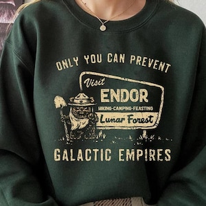 Puede incluir: Sudadera verde oscuro con el texto "ONLY YOU CAN PREVENT Visit ENDOR Hiking-Camping-Feasting Lunar Forest GALACTIC EMPIRES." El gráfico presenta un personaje de dibujos animados con una pala y un letrero.