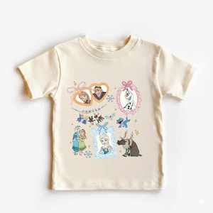 May include: A cream-coloured t-shirt featuring characters from the animated film "Frozen." The design includes framed images of Anna, Elsa, Olaf, and Kristoff, along with other characters and the word "FROZEN" in a decorative font.