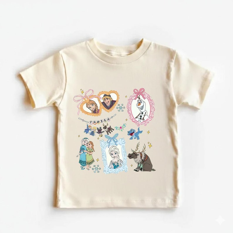 May include: A cream-coloured t-shirt featuring characters from the animated film "Frozen." The design includes framed images of Anna, Elsa, Olaf, and Kristoff, along with other characters and the word "FROZEN" in a decorative font.