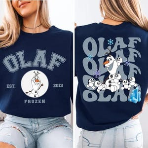 May include: Navy blue sweatshirt featuring the word "OLAF" in a collegiate font. The front design includes a circular graphic of Olaf the snowman from Frozen, with "EST. 2013" and "FROZEN" below. The back has a repeating "OLAF" design with Olaf and snowgies.
