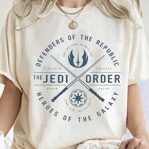 Può includere: T-shirt color crema con un design circolare. Il testo recita "Difensori della Repubblica" e "L'Ordine Jedi". Il design include spade laser incrociate, un emblema a stella e la frase "Che la Forza sia con te."