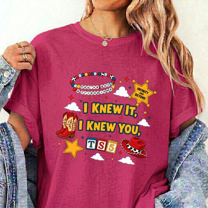 May include: A magenta t-shirt featuring a graphic print with a colorful beaded bracelet, a yellow sheriff star, a red cowboy boot, a red cowboy hat, and blocks spelling TS5. The text reads I KNEW IT, I KNEW YOU, and INFINITY AND BEYOND.