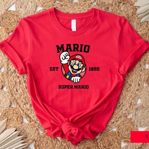 May include: Red t-shirt with a black graphic of Mario from the Super Mario video game series. The graphic includes the text "Mario Est 1985 Super Mario".