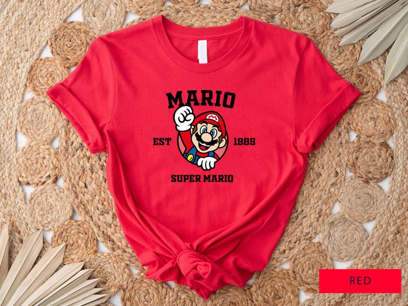 May include: Red t-shirt with a black graphic of Mario from the Super Mario video game series. The graphic includes the text "Mario Est 1985 Super Mario".