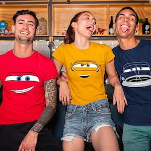 May include: Three people wearing t-shirts with cartoon car designs. The shirts are red, yellow, and navy blue. The red shirt has a smiling car face, the yellow shirt has a car face, and the navy blue shirt has a car design.