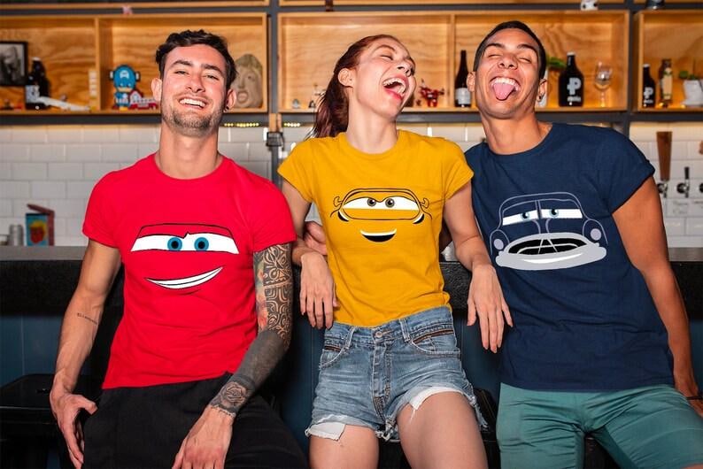 May include: Three people wearing t-shirts with cartoon car designs. The shirts are red, yellow, and navy blue. The red shirt has a smiling car face, the yellow shirt has a car face, and the navy blue shirt has a car design.