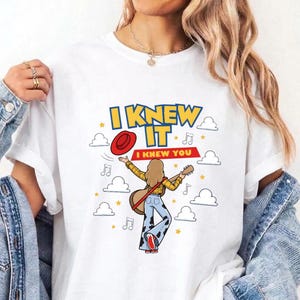 May include: A white t-shirt featuring a graphic of a person with long hair playing a guitar, wearing a yellow plaid shirt and cow-print pants. The text reads I KNEW IT and I KNEW YOU, surrounded by red hat, musical notes, stars, and cloud icons.