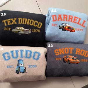 May include: Four sweatshirts with car-themed designs. A black sweatshirt features "TEX DINOCO EST. 1975" with a car graphic. A light blue sweatshirt displays "DARRELL EST. 1977". A beige sweatshirt has "GUIDO EST. 2006". A dark grey sweatshirt shows "SNOT ROD EST. 1968".