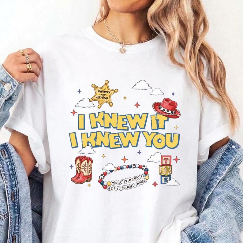Op de afbeelding: Een wit t-shirt met een grafische print met de tekst I KNEW IT I KNEW YOU in gele letters. Het ontwerp bevat een gouden sheriffster, een rode cowboyhoed, rode cowboylaarzen, een kleurrijke kralenarmband en gestapelde alfabetblokken.