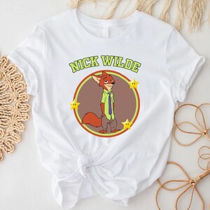 May include: White t-shirt featuring a cartoon illustration of Nick Wilde from Zootopia. The design includes a circular image of Nick Wilde, the text "NICK WILDE" above, and three yellow stars. The shirt is made of a soft material.