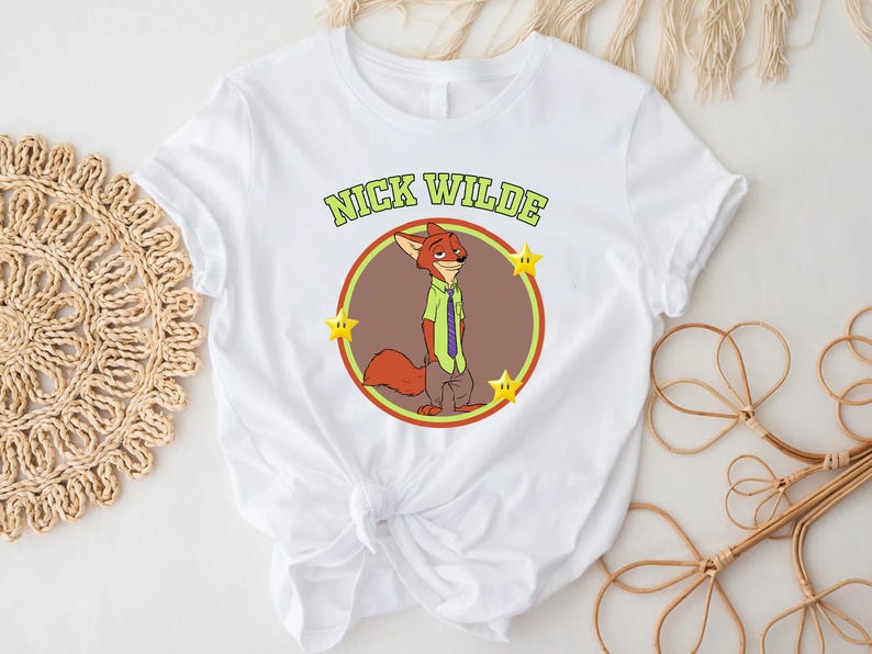May include: White t-shirt featuring a cartoon illustration of Nick Wilde from Zootopia. The design includes a circular image of Nick Wilde, the text "NICK WILDE" above, and three yellow stars. The shirt is made of a soft material.