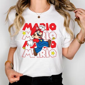 May include: A white short-sleeved t-shirt featuring a graphic of Mario jumping. The design includes the repeated red text MARIO in a bold font, along with yellow star and gold coin icons. The character wears a red hat, blue overalls, and brown shoes.