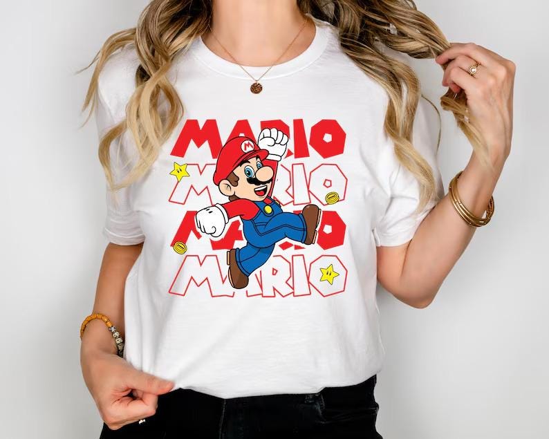 May include: A white short-sleeved t-shirt featuring a graphic of Mario jumping. The design includes the repeated red text MARIO in a bold font, along with yellow star and gold coin icons. The character wears a red hat, blue overalls, and brown shoes.
