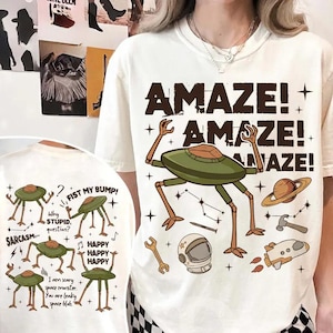May include: Off-white t-shirt with a large graphic featuring a green alien-like creature and the word "AMAZE!" repeated. Additional space-themed illustrations include a planet, rocket, and tools. The shirt also has smaller alien graphics.