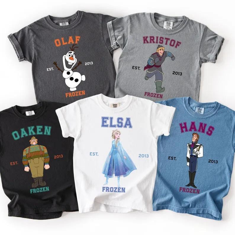 May include: Five short-sleeved t-shirts featuring characters from the film "Frozen." Each shirt displays a character's name in a collegiate font, along with an illustration and the word "FROZEN." Colours include black, white, grey, and blue.