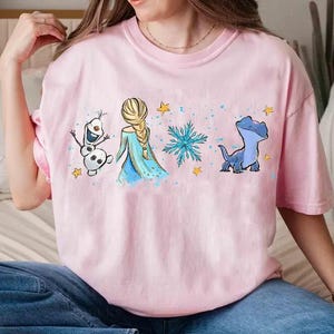 May include: A light pink t-shirt featuring a graphic of characters from a popular animated film. The design includes a blonde-haired woman in a blue dress, a snowman, a blue lizard, snowflakes, and yellow stars.