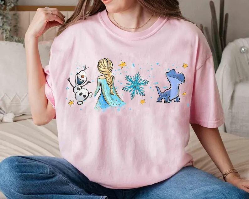 May include: A light pink t-shirt featuring a graphic of characters from a popular animated film. The design includes a blonde-haired woman in a blue dress, a snowman, a blue lizard, snowflakes, and yellow stars.