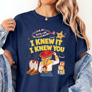 May include: A navy blue t-shirt featuring a graphic print with the text I KNEW IT I KNEW YOU, a beaded necklace, a yellow star, a pair of brown cowboy boots, a figure with blonde hair wearing a red hat, and three stacked alphabet blocks.