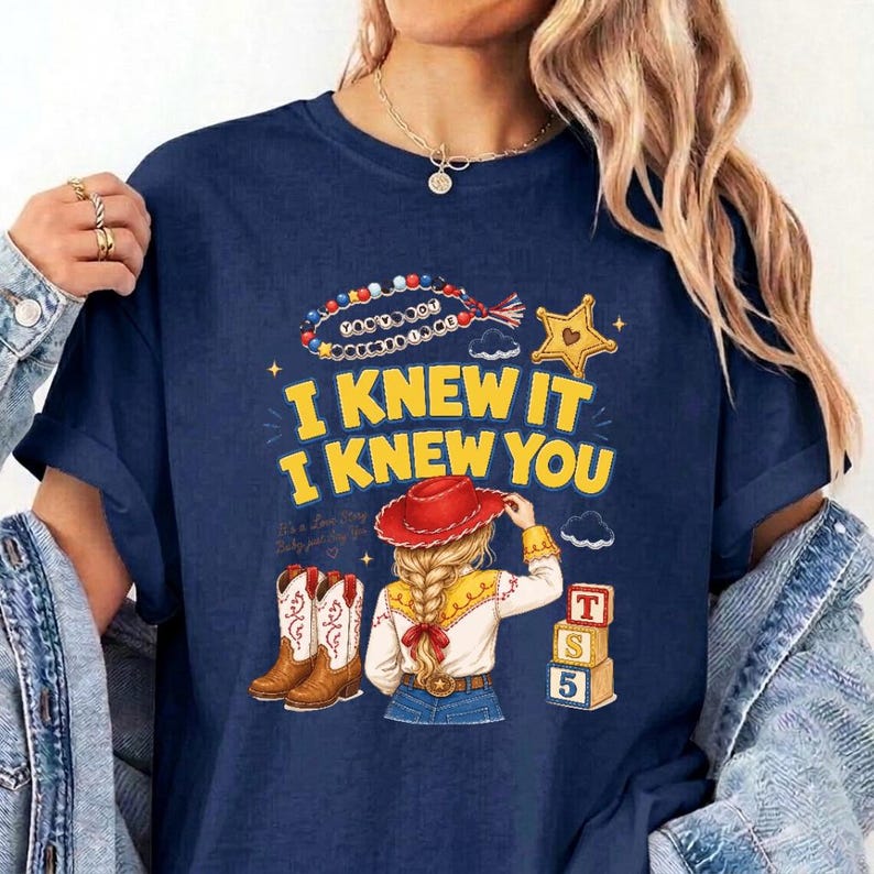 May include: A navy blue t-shirt featuring a graphic print with the text I KNEW IT I KNEW YOU, a beaded necklace, a yellow star, a pair of brown cowboy boots, a figure with blonde hair wearing a red hat, and three stacked alphabet blocks.