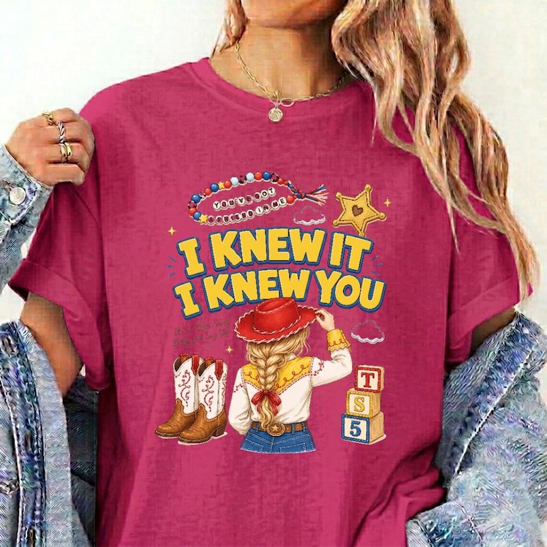 May include: A magenta t-shirt featuring a graphic print with the text I KNEW IT I KNEW YOU. The design includes a beaded string, a yellow star, a pair of brown cowboy boots, a blonde figure in a red hat, and a stack of three alphabet blocks.