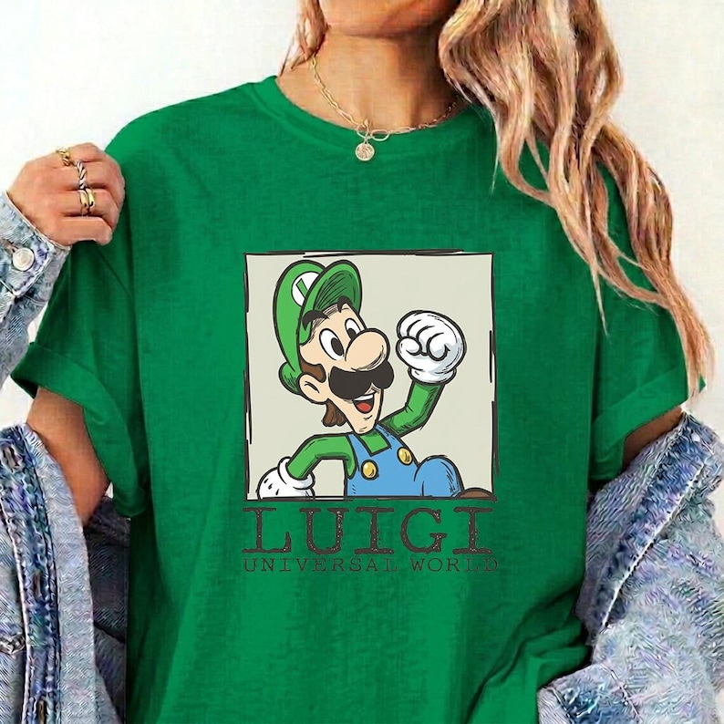 May include: A green short-sleeved t-shirt featuring a square graphic of the character Luigi with a mustache and green cap. Below the graphic, the text reads LUIGI UNIVERSAL WORLD in a dark, distressed font. The shirt has a crew neckline.