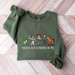 May include: An olive green sweatshirt featuring a graphic of cartoon characters and the text "YOU'VE GOT A FRIEND IN ME". The design includes a green dinosaur, Buzz Lightyear, Jessie, Woody, and a horse.