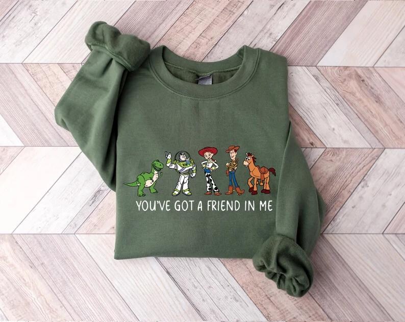 May include: An olive green sweatshirt featuring a graphic of cartoon characters and the text "YOU'VE GOT A FRIEND IN ME". The design includes a green dinosaur, Buzz Lightyear, Jessie, Woody, and a horse.