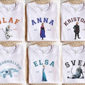 May include: White t-shirts with characters from the film Frozen. Each shirt displays a character's name in a varsity style font, with the year "2013" beneath. Characters include Olaf, Anna, Kristoff, Marshmallow, Elsa, and Sven.
