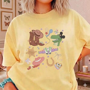 May include: Yellow t-shirt featuring a colorful graphic of cartoon cowboy-themed elements. The design includes a cowboy hat, boots, a sheriff badge, a dinosaur, a space ship, and other playful illustrations. The shirt is a comfortable, casual style.