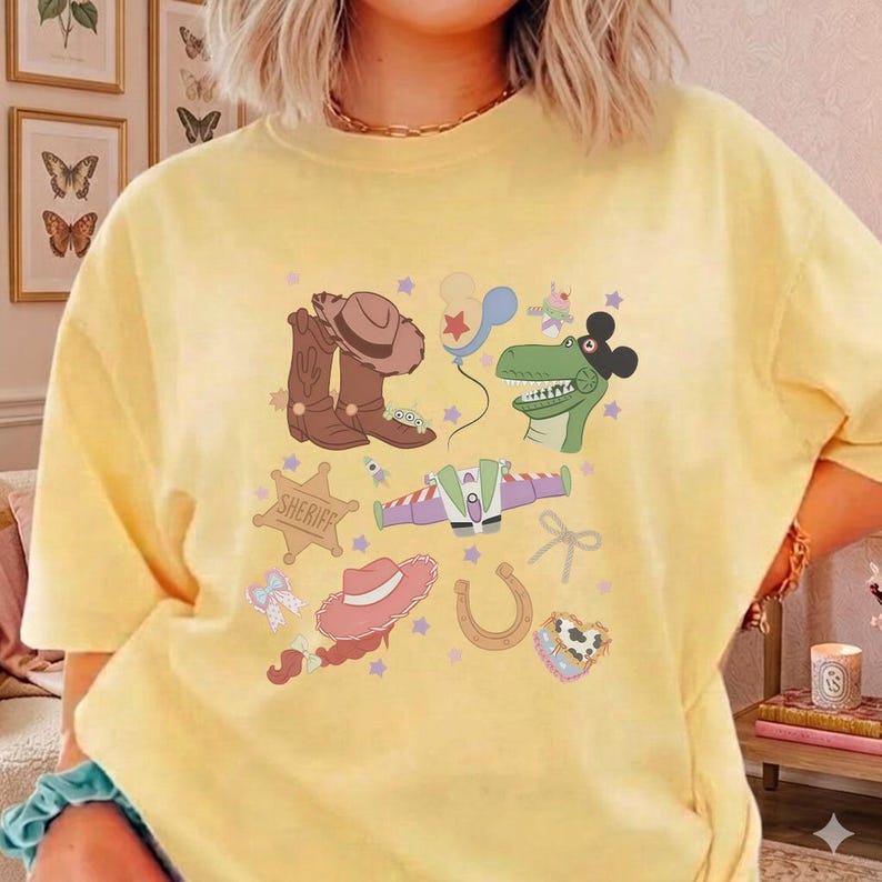 May include: Yellow t-shirt featuring a colorful graphic of cartoon cowboy-themed elements. The design includes a cowboy hat, boots, a sheriff badge, a dinosaur, a space ship, and other playful illustrations. The shirt is a comfortable, casual style.