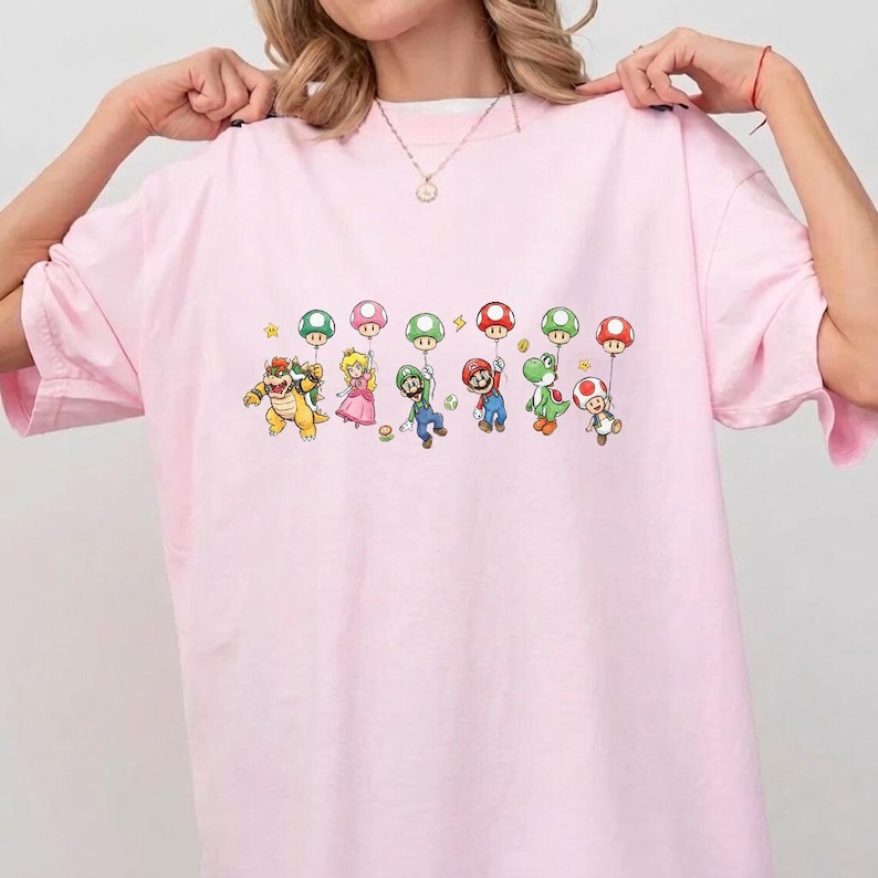 May include: A light pink short-sleeved t-shirt featuring a graphic print of various video game characters, including Bowser, Princess Peach, Luigi, Mario, Yoshi, and Toad, each holding a colorful mushroom-shaped balloon above their heads.