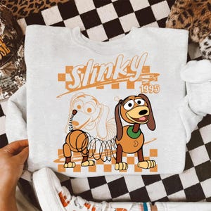 Może przedstawiać: Jasnoszara bluza z okrągłym dekoltem z nadrukiem graficznym przedstawiającym postać Slinky Dog. Wz&oacute;r zawiera napis Slinky 1995, pomarańczowe tło w kratkę oraz ilustrację konturową psa obok wersji kolorowej.