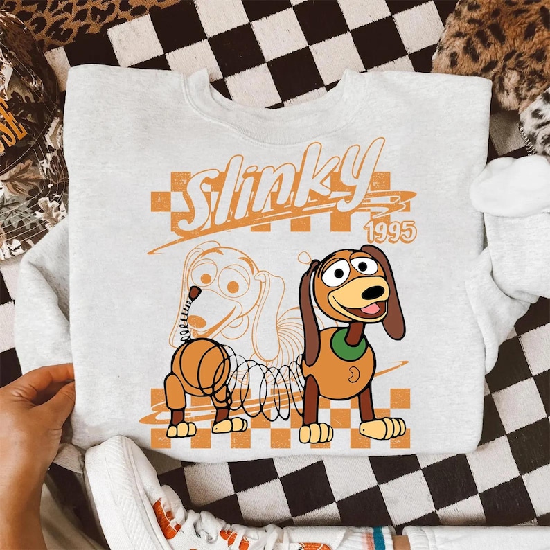 Może przedstawiać: Jasnoszara bluza z okrągłym dekoltem z nadrukiem graficznym przedstawiającym postać Slinky Dog. Wz&oacute;r zawiera napis Slinky 1995, pomarańczowe tło w kratkę oraz ilustrację konturową psa obok wersji kolorowej.
