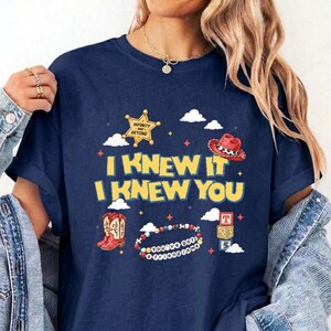 Op de afbeelding: Een marineblauw t-shirt met een grafische print met de tekst I KNEW IT I KNEW YOU. Het ontwerp bevat een gele sheriffster, een rode cowboyhoed, een kralenarmband met de tekst YOU'VE GOT A FRIEND IN ME en gestapelde alfabetblokken.