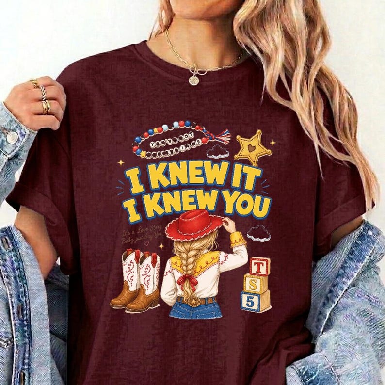 May include: A maroon t-shirt featuring a graphic print with the text I KNEW IT I KNEW YOU. The design includes a beaded string, a yellow star, a pair of brown cowboy boots, a person with blonde braided hair wearing a red hat, and two blocks.