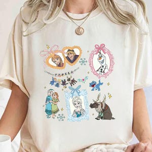 May include: Cream-coloured t-shirt with colourful illustrations of characters from the animated film "Frozen." The design includes framed images of Anna, Elsa, Kristoff, Olaf, and Sven, along with the word "FROZEN."