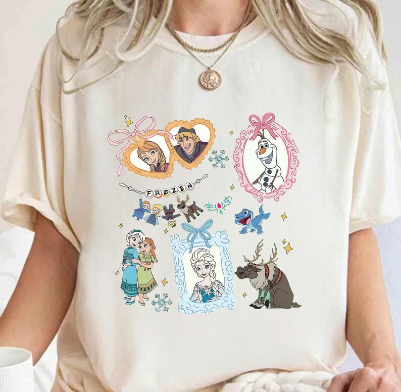 May include: Cream-coloured t-shirt with colourful illustrations of characters from the animated film "Frozen." The design includes framed images of Anna, Elsa, Kristoff, Olaf, and Sven, along with the word "FROZEN."