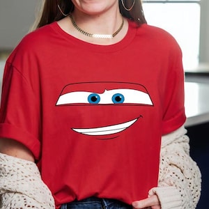 May include: Red t-shirt featuring a cartoon car face with blue eyes and a smiling mouth. The graphic is centered on the shirt. The shirt has short sleeves and a crew neck.