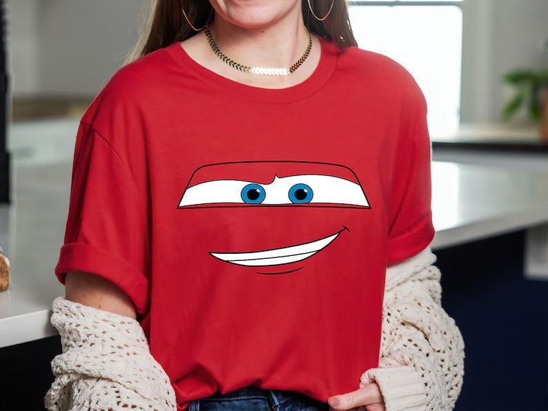 May include: Red t-shirt featuring a cartoon car face with blue eyes and a smiling mouth. The graphic is centered on the shirt. The shirt has short sleeves and a crew neck.