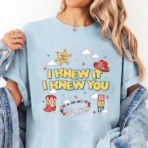 Op de afbeelding: Een lichtblauw t-shirt met een grafische print met de tekst I KNEW IT I KNEW YOU, een gele sheriffster, een rode cowboyhoed, rode cowboylaarzen, een kleurrijke kralenarmband met tekst en een stapel van drie blokken met letters.