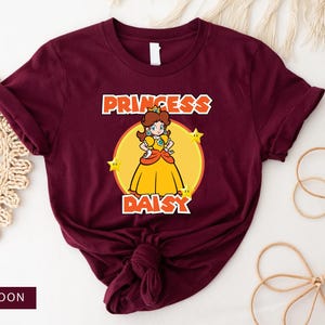 Könnte beinhalten: Ein maroonfarbenes T-Shirt mit einem Aufdruck von Prinzessin Daisy aus dem Super Mario-Franchise. Der Aufdruck zeigt Daisy in ihrem charakteristischen gelben Kleid mit einer Krone und Sternen. Der Text "Princess Daisy" ist in Orange unter dem Aufdruck gedruckt.