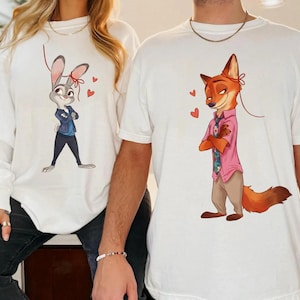 K&ouml;nnte beinhalten: Wei&szlig;es Langarm-Sweatshirt und T-Shirt mit Zeichentrickfiguren. Das Sweatshirt hat eine Judy Hopps-Grafik, und das T-Shirt hat eine Nick Wilde-Grafik. Beide Designs enthalten rote Herz-Akzente.