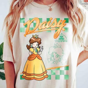 May include: A cream-coloured t-shirt with a cartoon illustration of Princess Daisy from the Super Mario franchise. The design includes the word "Daisy" in orange script, a green and orange checkered background, and a sketch of the character.