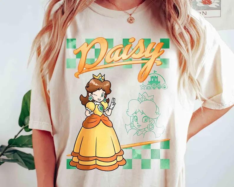 May include: A cream-coloured t-shirt with a cartoon illustration of Princess Daisy from the Super Mario franchise. The design includes the word "Daisy" in orange script, a green and orange checkered background, and a sketch of the character.