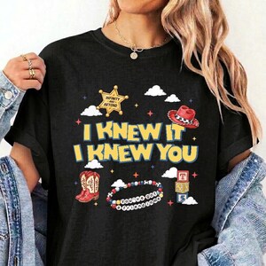 Puede incluir: Una camiseta negra con un estampado gr&aacute;fico que incluye el texto I KNEW IT I KNEW YOU, una estrella de sheriff amarilla, un sombrero de vaquero rojo, botas de vaquero rojas, una pulsera de cuentas colorida con texto y tres cubos apilados.