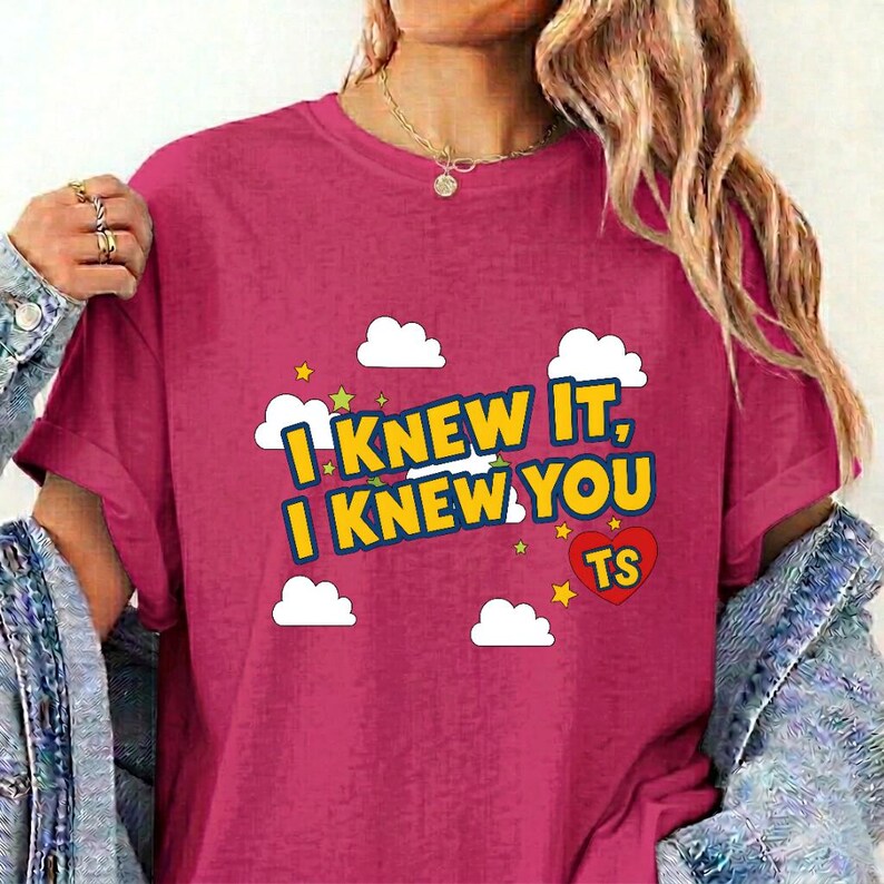 May include: A magenta short-sleeved t-shirt featuring a graphic print of white clouds, small yellow stars, and a red heart with the letters TS inside. The text reads I KNEW IT, I KNEW YOU in bold yellow letters with a dark blue outline on the front.