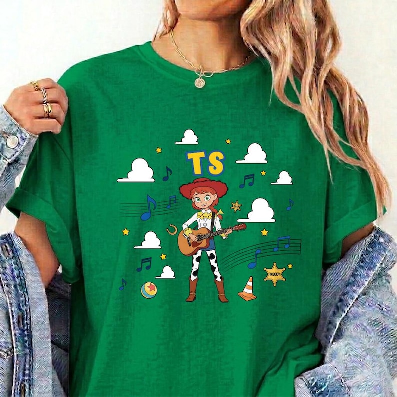 May include: A green t-shirt featuring a graphic of a cartoon cowgirl playing an acoustic guitar. The design includes the yellow text TS, white clouds, blue musical notes, yellow stars, a sheriff badge, a ball, and an orange traffic cone.